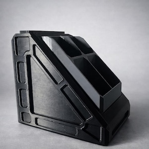 May include: A black, angled desk organizer featuring a triangular side profile with recessed rectangular panels and a top section divided into four distinct compartments for storage. The structure is solid and features a clean, geometric design.