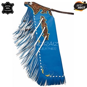 Puede incluir: Chaps de cuero azul brillante con largos flecos blancos y azules en el lateral. La sección superior muestra un intrincado grabado floral marrón con cordones blancos. Una correa de cuero marrón con hebilla está unida en la parte superior. Texto NORAIZ LEATHER.