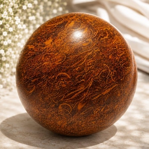 Puede incluir: Una esfera decorativa de madera, de color marrón oscuro, con intrincados diseños en espiral grabados en su superficie. Presenta detalles en marrón claro y se encuentra sobre una superficie de tono neutro.