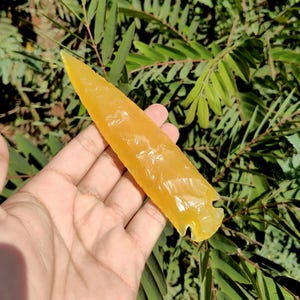 May include: A hand holding a translucent, yellow-orange arrowhead. The arrowhead is elongated with a sharp point and a notched base. The background features green foliage.