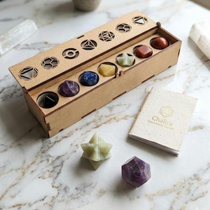 Op de afbeelding: Een houten doos met een scharnierend deksel, met zeven kleurrijke edelstenen. Het deksel heeft geometrische uitsparingen. Een klein boekje met de titel "Chakra Geometry Guide" ligt naast de doos. Twee extra edelstenen zijn ook zichtbaar.