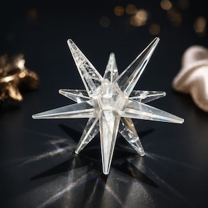 May include: A clear, multi-pointed star-shaped decorative object. The star has sharp, angular points and a faceted surface, creating a sparkling effect. The object is likely made of crystal or glass, and is set against a dark background.