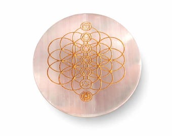 May include: A circular, pale pink selenite crystal plate with a gold Sacred Geometry design. The design features overlapping circles and symbols. The plate is approximately 10 cm in diameter.
