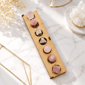 Puede incluir: Un soporte de exhibición rectangular de madera con siete aberturas circulares, cada una con una forma de cristal geométrica única. La colección incluye varias piedras de cuarzo rosa y cuarzo transparente en formas como esferas, pirámides y cubos.