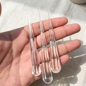 Könnte beinhalten: Klare, tropfenförmige Glasanhänger mit spitzen Spitzen. Die Anhänger werden in einer Hand gehalten, was ihre transparente Qualität und ihr längliches Design hervorhebt. Das Glas wirkt glatt und reflektierend und fängt das Licht ein.