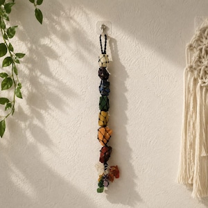 Può includere: Una decorazione da parete verticale con sette pietre grezze multicolori racchiuse in una rete di macramè nera. Le pietre sono disposte in colonna e presentano tonalità bianche, viola, blu, verdi, gialle, arancioni e rosse, terminando con cristalli.