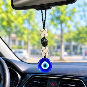 Könnte beinhalten: Ein hängendes Ornament für den Rückspiegel mit einem kreisförmigen blauen Glas-Amulett in Form eines bösen Blicks, das an einer schwarzen Kordel hängt. Es enthält zwei weiße Würfelperlen und eine schwarze, strukturierte Perle in einem schwarzen Netz.