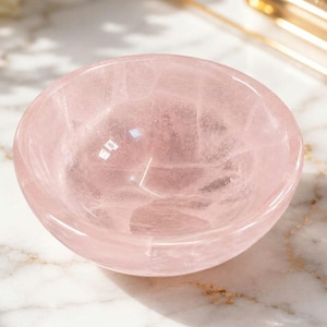 May include: A small, round bowl carved from translucent pale pink rose quartz. The stone features a smooth, polished surface with visible natural internal fractures and crystalline patterns throughout the material, creating a soft, glossy appearance.