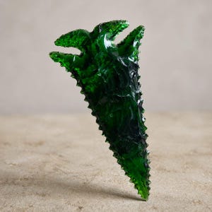 May include: A dark green, triangular glass sculpture with a jagged edge and three upward-pointing prongs at the top. The artwork has a textured surface and is displayed against a neutral background.