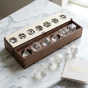 Könnte beinhalten: Ein Holzbox-Set mit geometrischen Formen aus klarem Quarz-Kristall. Die Box hat einen hellen Deckel mit geometrischen Ausschnitten. Das Set enthält eine Pyramide, eine Kugel, einen Würfel und andere Formen. Ein kleines Büchlein mit goldenem Text ist ebenfalls enthalten.