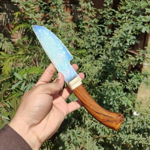 Puede incluir: Un cuchillo artesanal con una hoja opalina azul claro y un mango de madera. El mango es de color marrón cálido y está envuelto con una fibra natural cerca de la hoja. El cuchillo se muestra sobre un fondo de follaje verde.