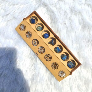 May include: A wooden box with geometric cutouts and compartments holding various blue gemstones. The box is rectangular with a hinged lid and is displayed on a white, fluffy surface. The gemstones are of different shapes and sizes.