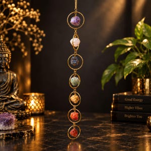 May include: A vertical hanging ornament featuring seven raw, multi-colored gemstones encased in gold-toned metal rings. The stones are arranged in a column, displaying shades of purple, pink, blue, green, yellow, orange, and red in a chain.