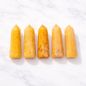 Op de afbeelding: Vijf gele kwartskristalpunten in een rij. De kristallen hebben een gepolijst, glad oppervlak en een taps toelopende, rechthoekige vorm. De kleur varieert van lichtgeel tot een diepere, gouden tint.