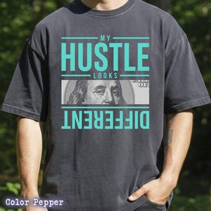May include: Dark gray t-shirt with the text "MY HUSTLE LOOKS DIFFERENT" in teal. The word "HUSTLE" is above a graphic of a $100 bill. The shirt is a relaxed fit.