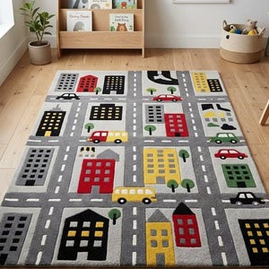 May include: A rectangular area rug featuring a city street grid pattern with gray roads, white lane markings, and various multi-story buildings in red, yellow, black, and gray. The design includes small colorful cars and green trees throughout.
