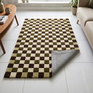 Op de afbeelding: Een rechthoekig vloerkleed met een dambordpatroon in bruin-, groen- en cr&egrave;metinten. Het kleed is gedeeltelijk opgerold en onthult een grijze achterkant. Het kleed ligt op een witte tegelvloer.