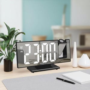 以下が含まれることがあります： 白数字で23:00を表示する黒いデジタル目覚まし時計。時計には投影機能があり、植物、メモ帳、ペンが置かれた机の上にあります。時計のディスプレイは長方形で、鏡面になっています。