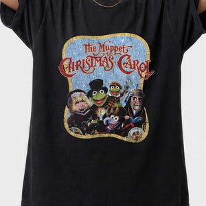 Puede incluir: Camiseta negra con un gráfico de "The Muppet Christmas Carol". El diseño incluye el título de la película y varios personajes de los Muppets. El gráfico está sobre un fondo azul con un borde dorado.