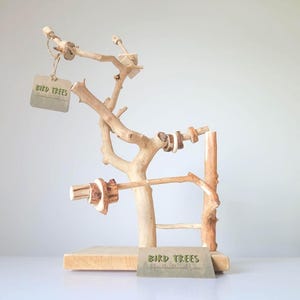 May include: A natural wood bird perch stand featuring multiple branches and a flat rectangular base. The structure includes several small wooden rings attached to the branches and two tan rectangular tags with the text BIRD TREES printed on them.