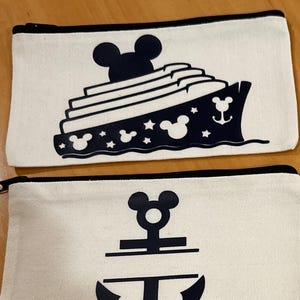 May include: A collection of zippered pouches. Three white pouches feature navy blue designs: a cruise ship, and two anchor designs. A black pouch displays a gold silhouette of a fairy with a wand and stars. All pouches have black zippers.