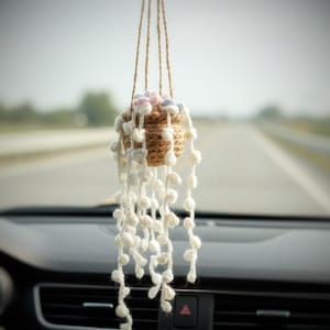 May include: A handmade crochet hanging plant basket in a car. The basket is brown with white, pink, and blue accents, and white beaded strands. The plant is suspended from the rearview mirror by brown twine.