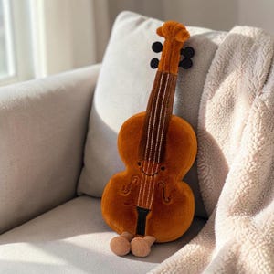 Puede incluir: Un juguete de violín naranja de peluche con una cara sonriente, detalles negros y pies beige. El juguete está sobre un sofá gris claro con una manta esponjosa de color crema. El violín tiene detalles de cuerdas blancas.