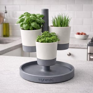 Può includere: Una fioriera per erbe aromatiche a tre livelli con tre vasi bianchi e grigi pieni di erbe fresche. La fioriera poggia su una base grigia con la scritta "wynetec" stampata sopra. L'ambiente della cucina comprende un lavandino e un fornello.
