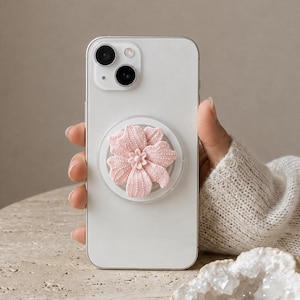 May include: A white smartphone featuring a circular, light pink, textured flower-shaped grip attached to the back. The flower has five distinct petals with a dotted surface pattern and a small central bud, centered on the back of the device.