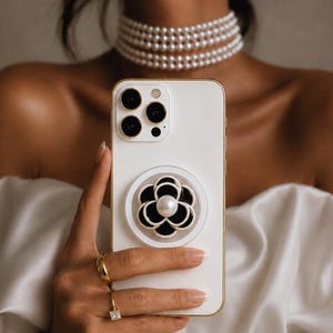 Pode incluir: Uma capa de smartphone branca com um suporte circular apresentando um design de flor de camélia em preto e branco com uma pérola central. O telefone possui um sistema de câmara tripla na parte traseira e uma borda dourada.