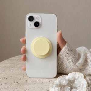 Op de afbeelding: Een witte smartphone met een dubbel camerasysteem aan de achterzijde en een ronde, lichtgele inklapbare telefoongreep in het midden. Het apparaat heeft een gladde, matte afwerking op de greep en een glanzende witte behuizing op de telefoon.