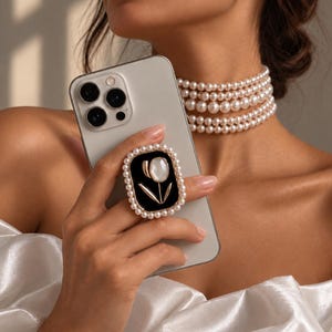 Peut inclure: Un support de téléphone rectangulaire avec un centre noir orné d'une tulipe dorée et d'une fleur nacrée blanche. Le support est entouré d'un cadre de petites perles blanches artificielles et est fixé au dos d'un smartphone de couleur claire.