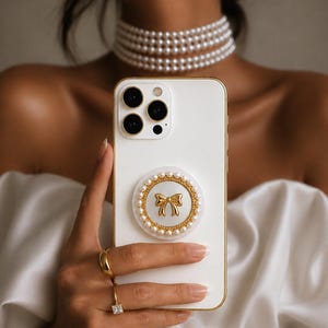 May include: A white smartphone case featuring a circular grip attachment with a gold-toned bow emblem at the center, surrounded by a border of small white pearls. The case has a gold-colored edge and a triple-lens camera cutout on the back.