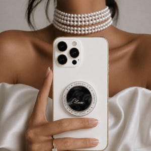 Pode incluir: Um smartphone branco com um suporte circular na parte traseira. O suporte tem um centro preto marmorizado com a palavra Love em letra cursiva branca, rodeado por uma borda prateada com o texto repetitivo Gift For You e pequenos pontos.