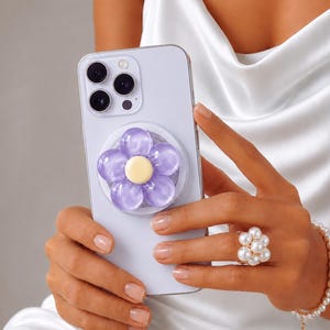 Peut inclure: Un smartphone violet clair doté d'un système de caméra à trois objectifs au dos. Au centre du téléphone est fixé un support circulaire en forme de fleur avec des pétales violets translucides et un centre circulaire jaune pâle uni.