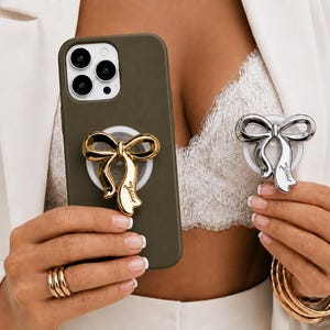 Pode incluir: Uma capa de smartphone verde-oliva com um suporte de telefone em forma de laço dourado fixado na parte traseira, ao lado de um suporte de telefone em forma de laço prateado na outra mão, ambos com acabamento metálico e design de fita.
