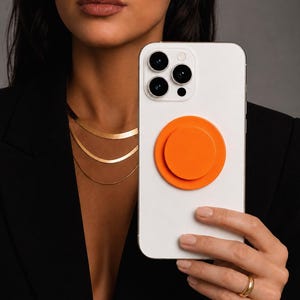 Op de afbeelding: Een witte smartphone met een ronde, massieve oranje inklapbare grip aan de achterkant. Het apparaat heeft een camerasysteem met drie lenzen in een vierkante module. De telefoon wordt vastgehouden voor een zwarte blazer en gelaagde gouden kettingen.