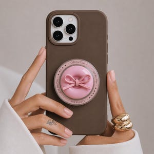 Peut inclure: Une coque de smartphone marron mat avec un support de téléphone circulaire rose. L'accessoire présente un nœud rose au centre et une bordure avec le texte répété Gift For You et des petites étoiles imprimés en foncé sur un anneau rose clair.