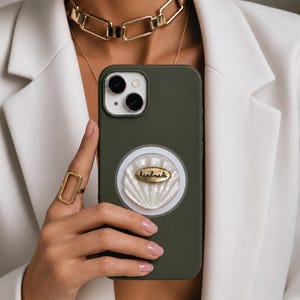 Pode incluir: Uma capa de smartphone verde-oliva fosca com um suporte circular branco em forma de concha na parte traseira. O centro da concha possui uma pequena placa de metal oval dourada com a palavra "handmade" gravada em letras pretas.