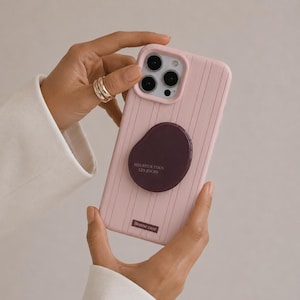 May include: A light pink phone case with a vertical ribbed texture, featuring a dark purple circular grip attachment printed with the text HEUREUX TOUS LES JOURS and a small rectangular label at the bottom with the text Bonne case.