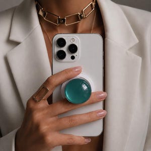 May include: A white smartphone featuring a circular, translucent teal phone grip attached to the back. The device is held against a white blazer, with a gold rectangular ring on a finger and a gold chain-link choker necklace visible.