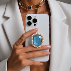 Peut inclure: Un smartphone blanc doté d'un support de téléphone décoratif à l'arrière. Le support a la forme d'une pierre précieuse bleue à facettes, entourée d'une bordure de petites perles blanches et d'un cadre en métal doré fixé au centre.