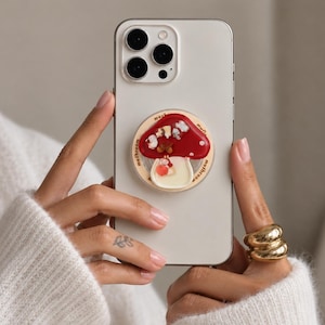 May include: A white smartphone featuring a circular phone grip on the back. The grip displays a red mushroom illustration with white spots, small decorative charms, and the word mushroom printed in a repeating pattern around the circular edge.