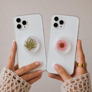 May include: Two white smartphones featuring circular phone grips on the back. One grip contains a pressed green fern leaf, while the other grip features a pressed pink flower with a brown centre, both encased in clear resin material.