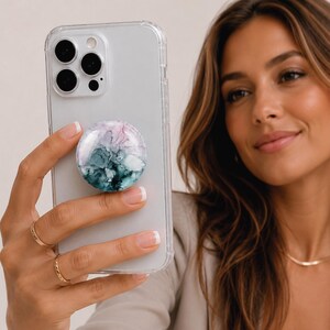 May include: A clear phone case with a colourful abstract design phone grip. The phone grip features shades of teal, purple, and white. The phone is held by a person.