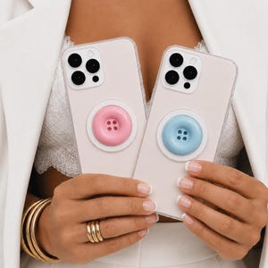 May include: Two smartphones with clear cases, each featuring a circular, button-shaped grip on the back. One grip is pastel pink and the other is pastel blue. Both phones have a triple-lens camera array in the upper left corner and a white base.