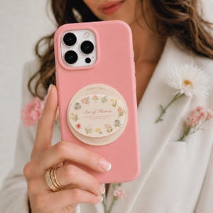 Puede incluir: Una funda de smartphone de color rosa sólido con un agarre circular plegable de color crema en la parte posterior. El agarre muestra un patrón floral que rodea el texto Sea of Flowers en letra cursiva, con pequeñas ilustraciones botánicas.