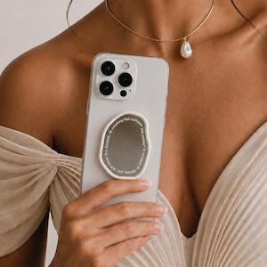 以下が含まれることがあります： 明るい色のスマートフォン。中央には不規則な形のクリーム色の円形グリップがあり、反射する鏡面と内側の縁に沿って小さな白い文字が印刷されています。オフホワイトの布地でドレープが施された衣服を背景にしています。