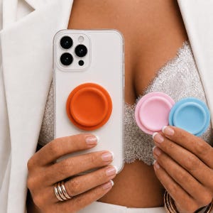 Könnte beinhalten: Eine transparente Smartphone-Hülle mit einem runden, orangen, einklappbaren Griff auf der Rückseite. Zwei weitere runde, einklappbare Griffe in Pastellrosa und Hellblau werden separat gehalten und zeigen eine glatte, matte Oberfläche.