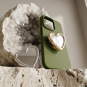 May include: An olive green phone case with a gold-rimmed heart-shaped phone grip. A silver heart-shaped compact mirror is placed beside the phone case. The background features a geode and a textured surface.